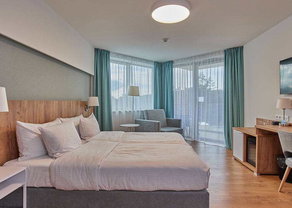 Nowoczesny pokój dwuosobowy z łóżkiem king, drewnianym zagłówkiem, lampkami nocnymi, fotelem, biurkiem, TV, minibarem i balkonem z woalem.