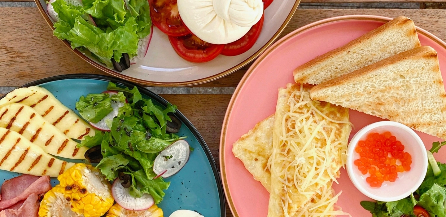 Plates on wooden table: omelette with grated cheese and toast, bowl of red salmon roe, salads, grilled cheese and corn.
