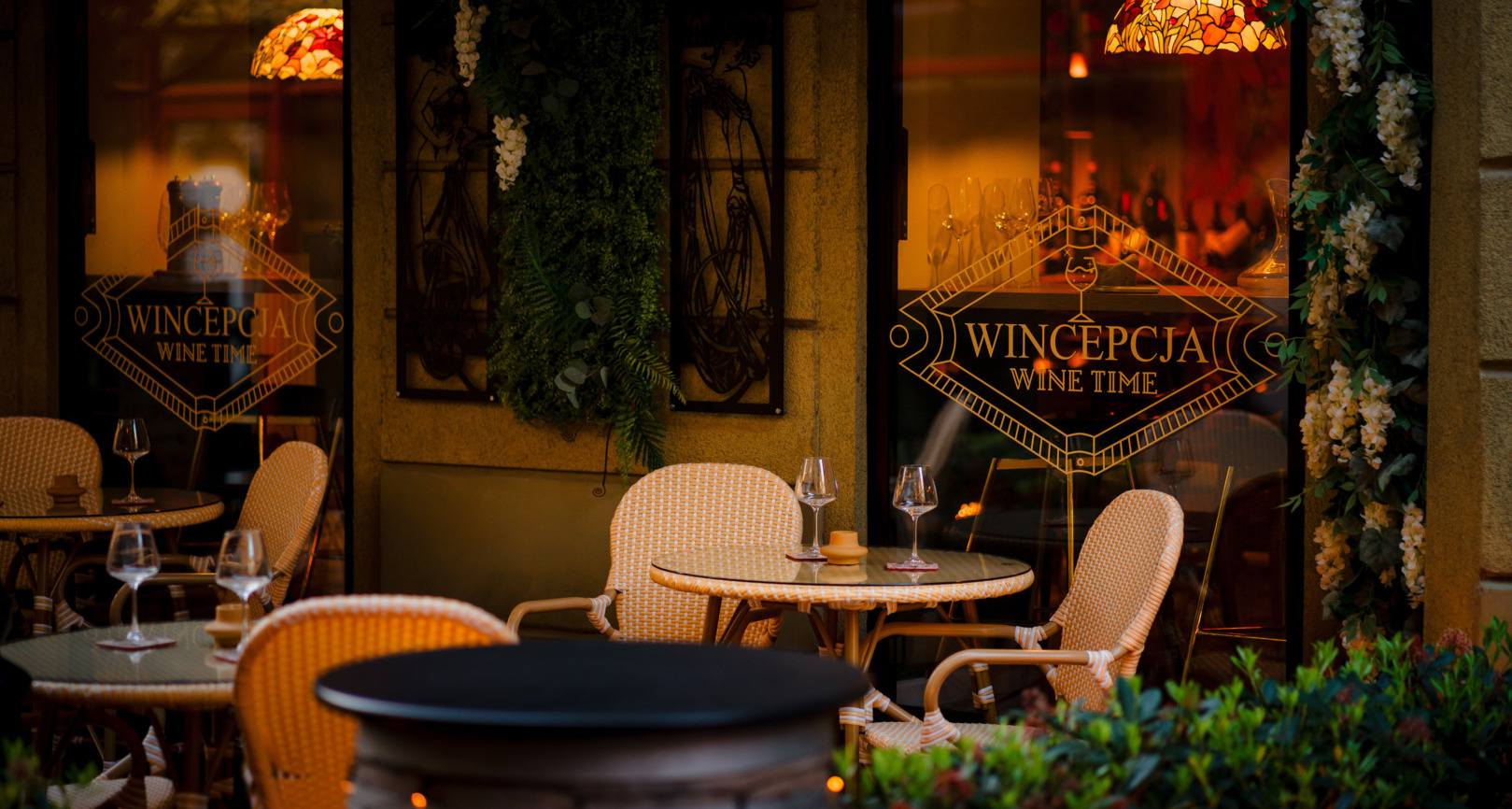 Outdoor seating area at a wine bar with rattan chairs, glass tables, wine glasses, and warm ambient lighting.