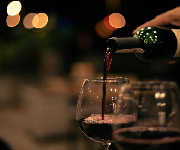 A hand pours red wine into a glass at a dimly lit table, while blurred lights create an intimate restaurant atmosphere