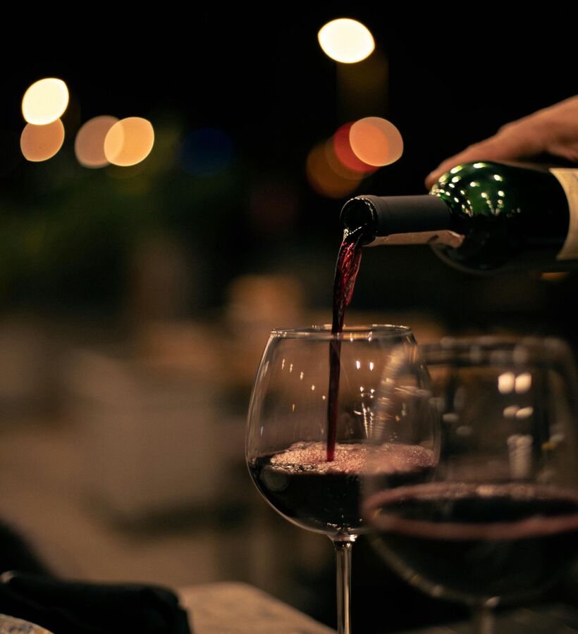 Hand pouring red wine into a stemmed glass at a dimly lit dining table, warm bokeh lights set an intimate restaurant mood