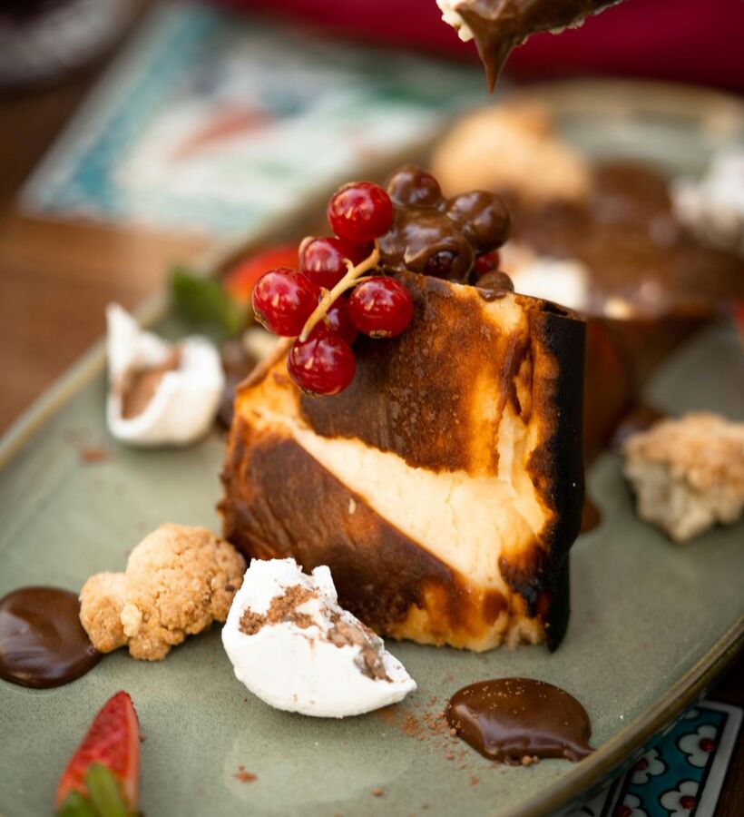 Charred-top cheesecake slice on a plate garnished with red currants, warm chocolate drizzle, meringue pieces and crumble