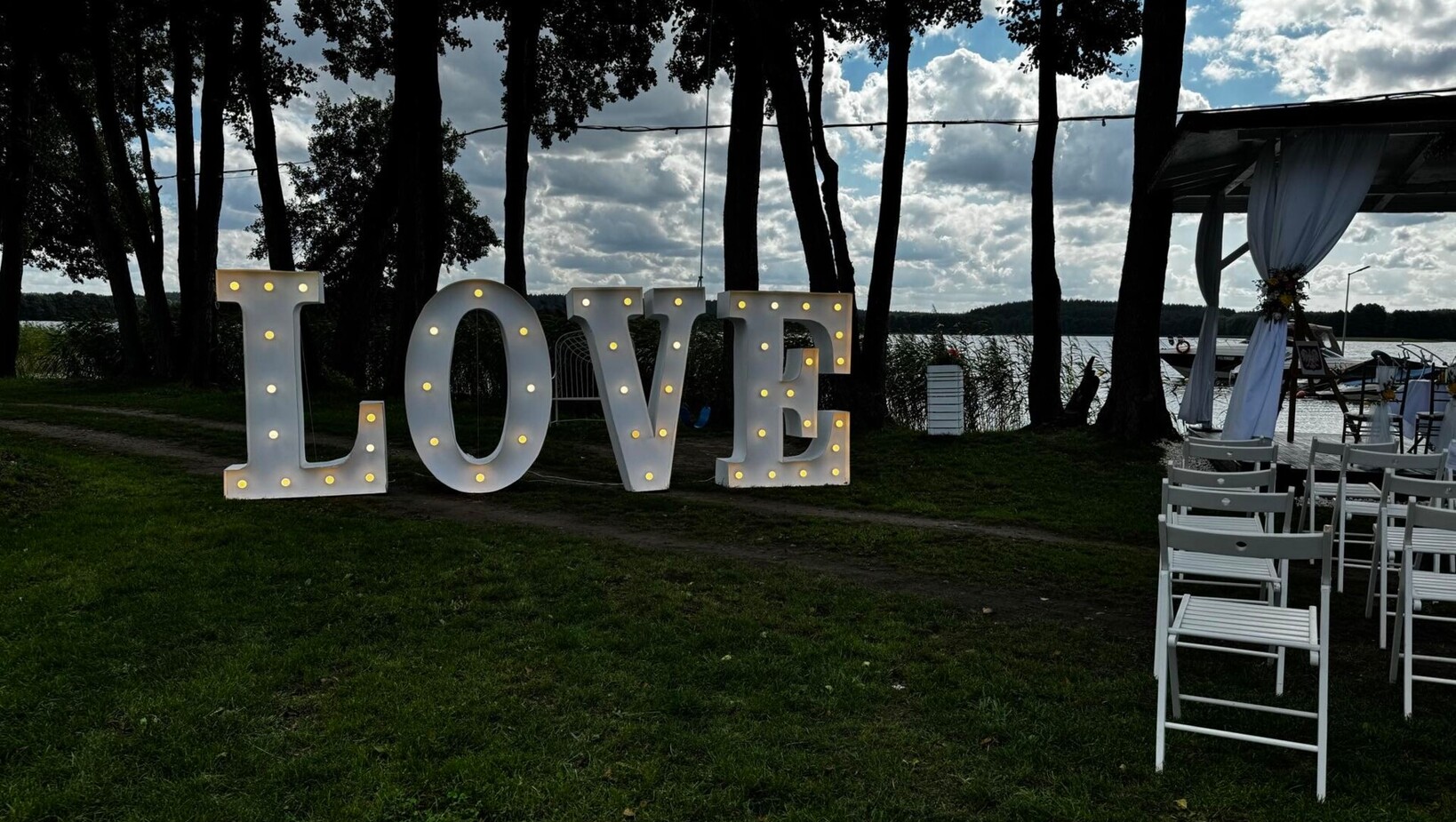 Duże podświetlane litery „LOVE” nad jeziorem pod drzewami, białe krzesła i przystrojona altana na pomoście na ceremonię.