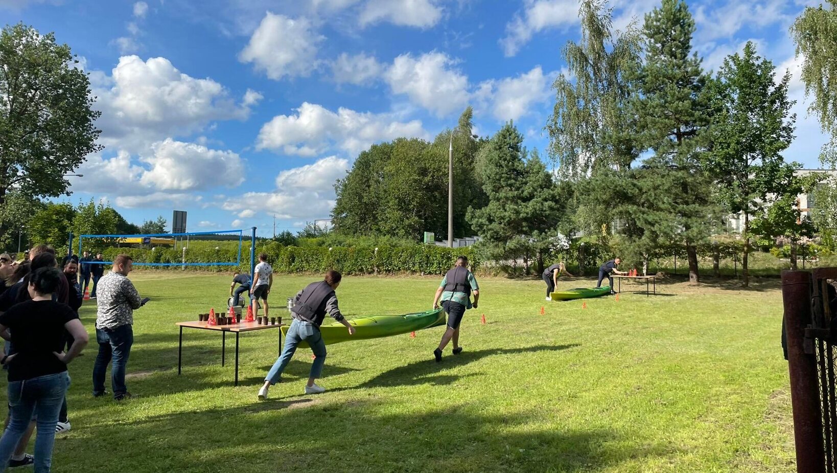 Teams carrying green kayaks across a sunny lawn during an outdoor team-building challenge, tables with cones nearby