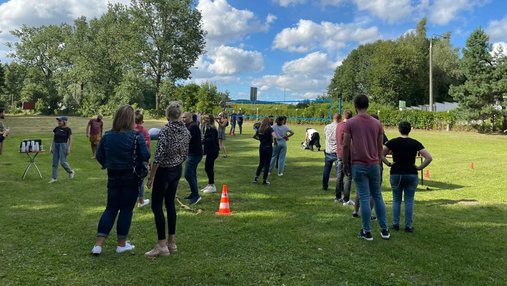Hotel guests at a sunny lawn event with volleyball net, cones and refreshment table amid mature trees and open grassy grounds