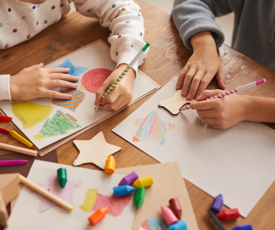 Two children coloring holiday shapes and decorating wooden star ornaments with crayons and colored pencils at a table