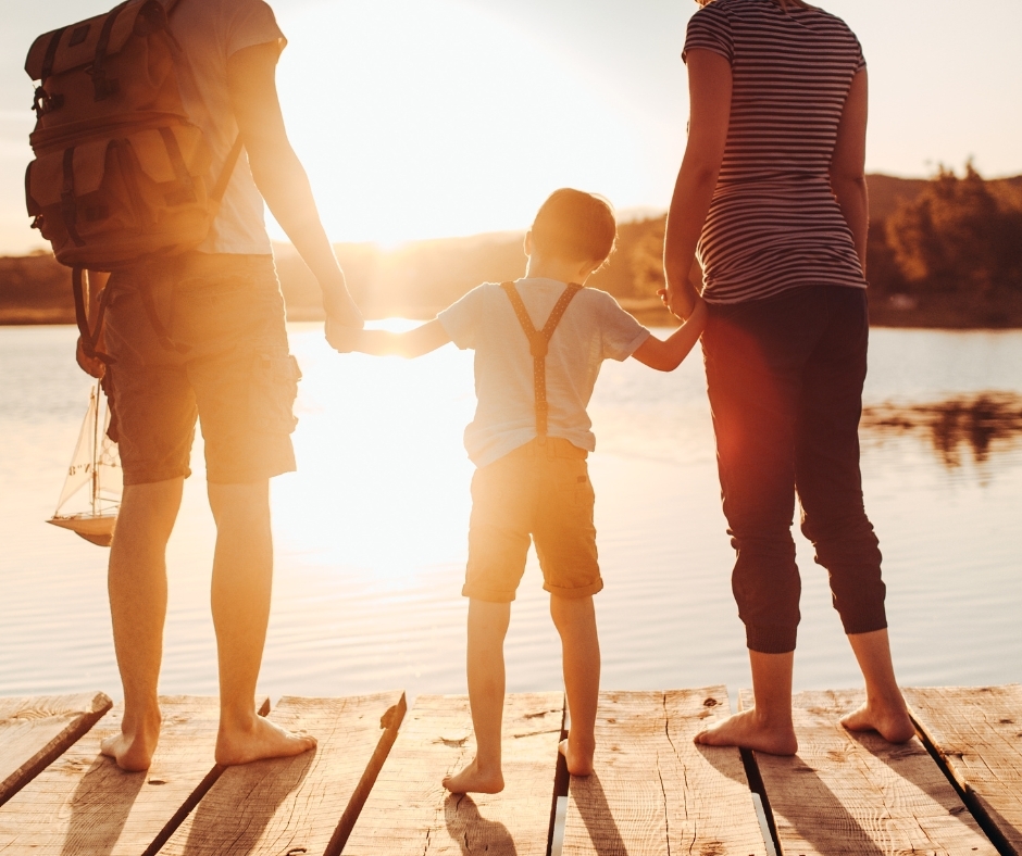 Family holding hands on a wooden dock at sunset by a calm lake, barefoot in warm golden light