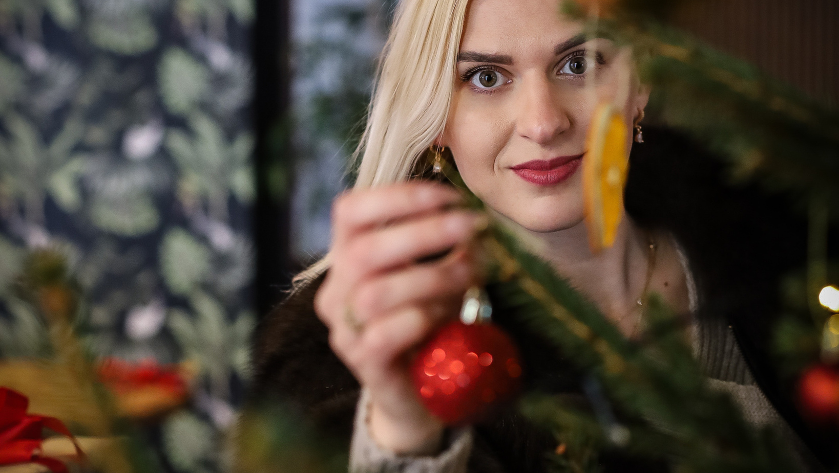 Woman hanging a red bauble on a decorated Christmas tree in a cozy, warmly lit hotel lobby with festive atmosphere