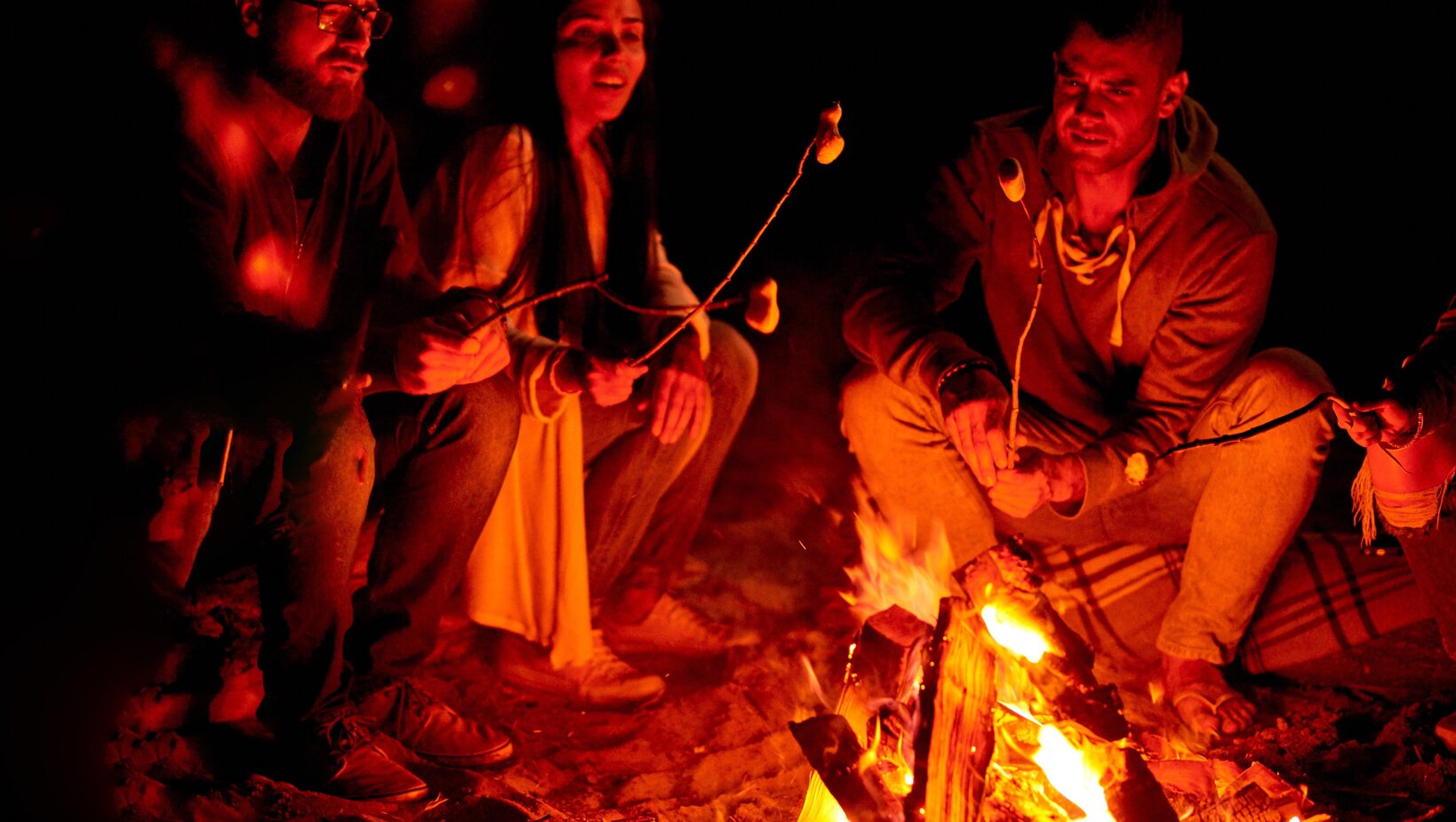 Group roasting marshmallows over a warm beach bonfire, cozy nighttime gathering by the fire