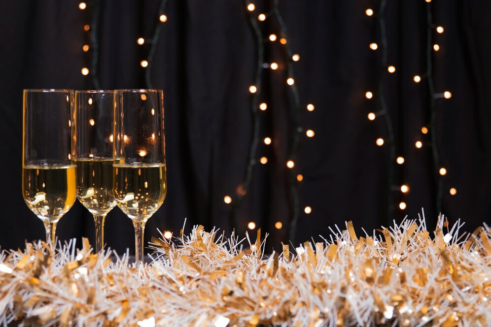 Three champagne flutes with sparkling wine on gold-and-white tinsel, warm fairy lights in background evoking a festive hotel celebration.