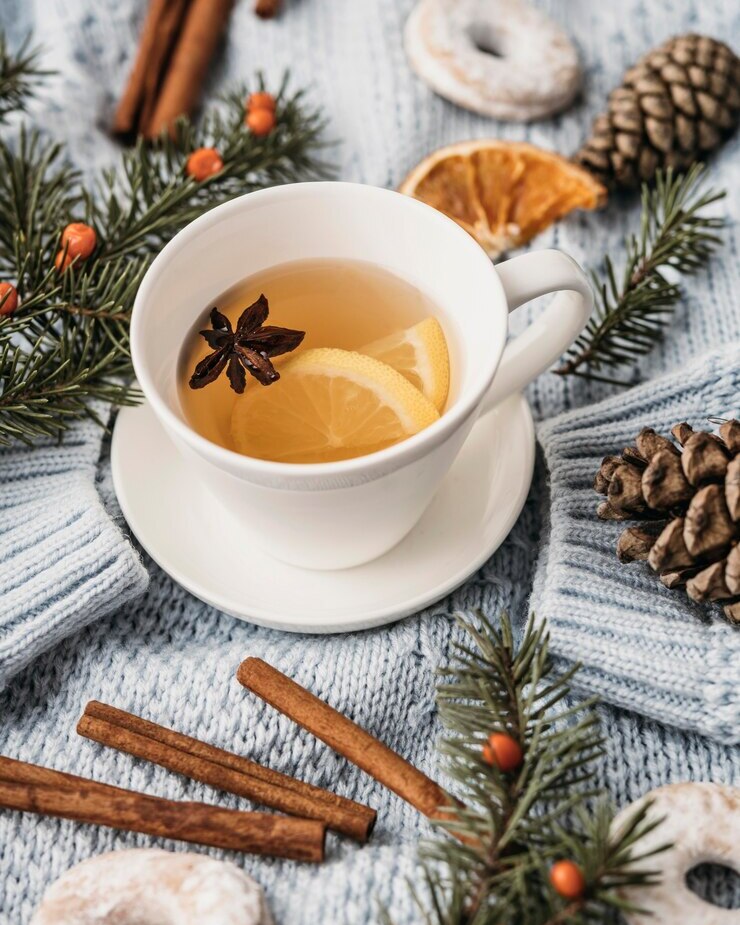 Weiße Tasse Zitronentee mit Sternanis auf blauer Decke, umgeben von Zimtstangen, Kiefernzweigen, Zapfen und Donuts