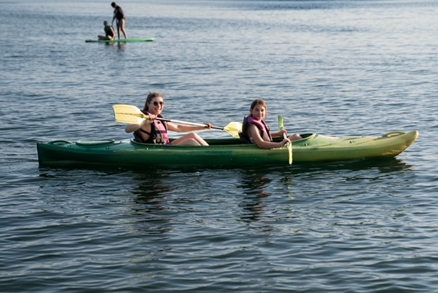 Two people paddling a green tandem kayak on calm water, wearing life jackets, sunlit recreational watersport