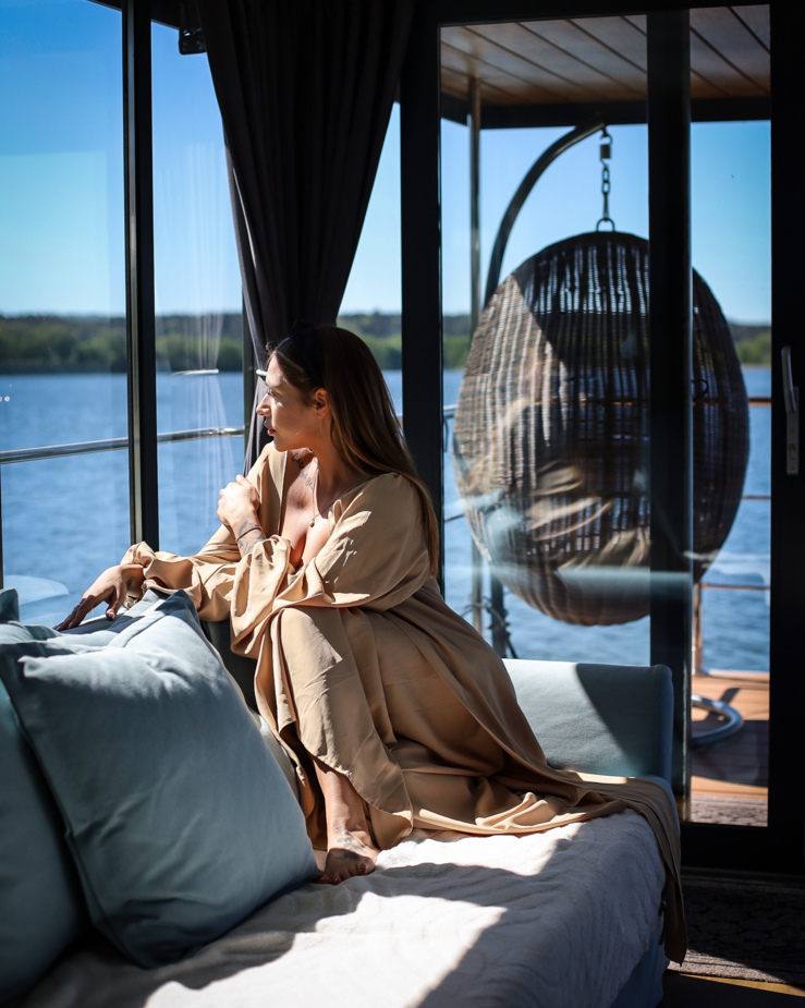 Frau im Bademantel entspannt auf dem Sofa einer Wasserblick-Suite, blickt durch große Fenster auf See und Terrasse mit Korbsessel