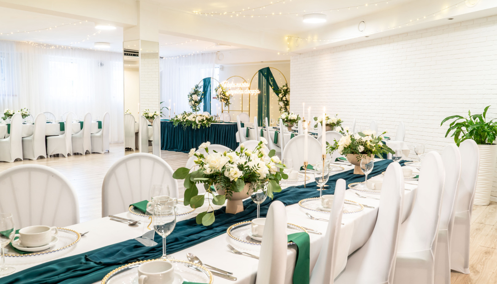 Elegant banquet room set for a wedding: long tables with white chair covers, teal runners, floral centerpieces, head‑table arch and string lights