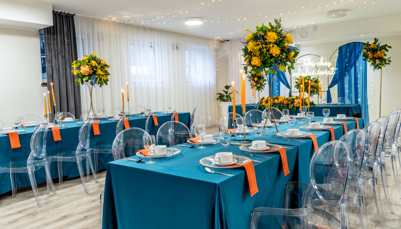 Elegant banquet room with teal tablecloths, orange napkins, clear ghost chairs, tall yellow floral centerpieces, candles and fairy-light backdrop