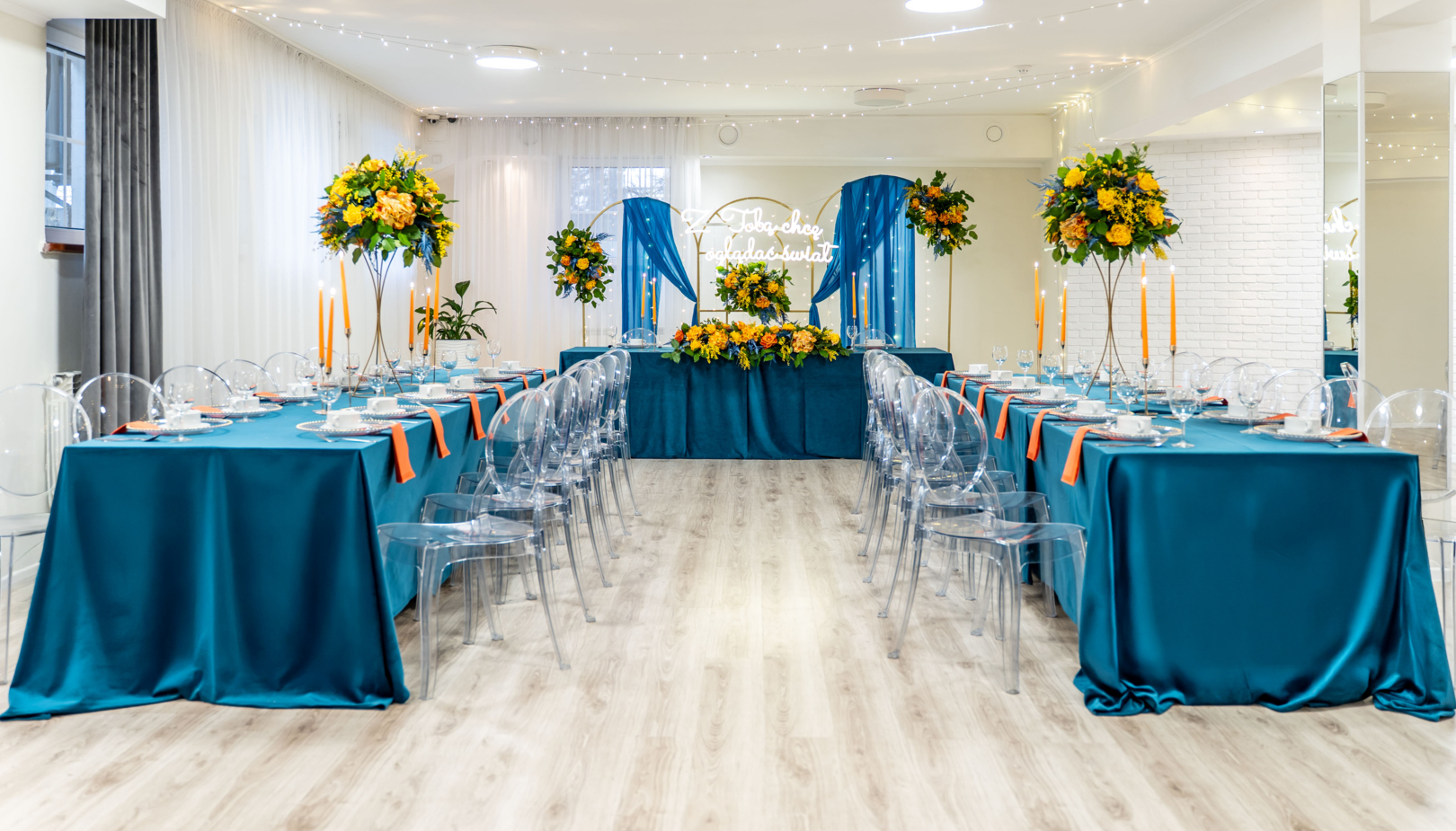 Banquet room with teal tablecloths, transparent chairs, tall yellow-orange floral centerpieces, candles and fairy lights
