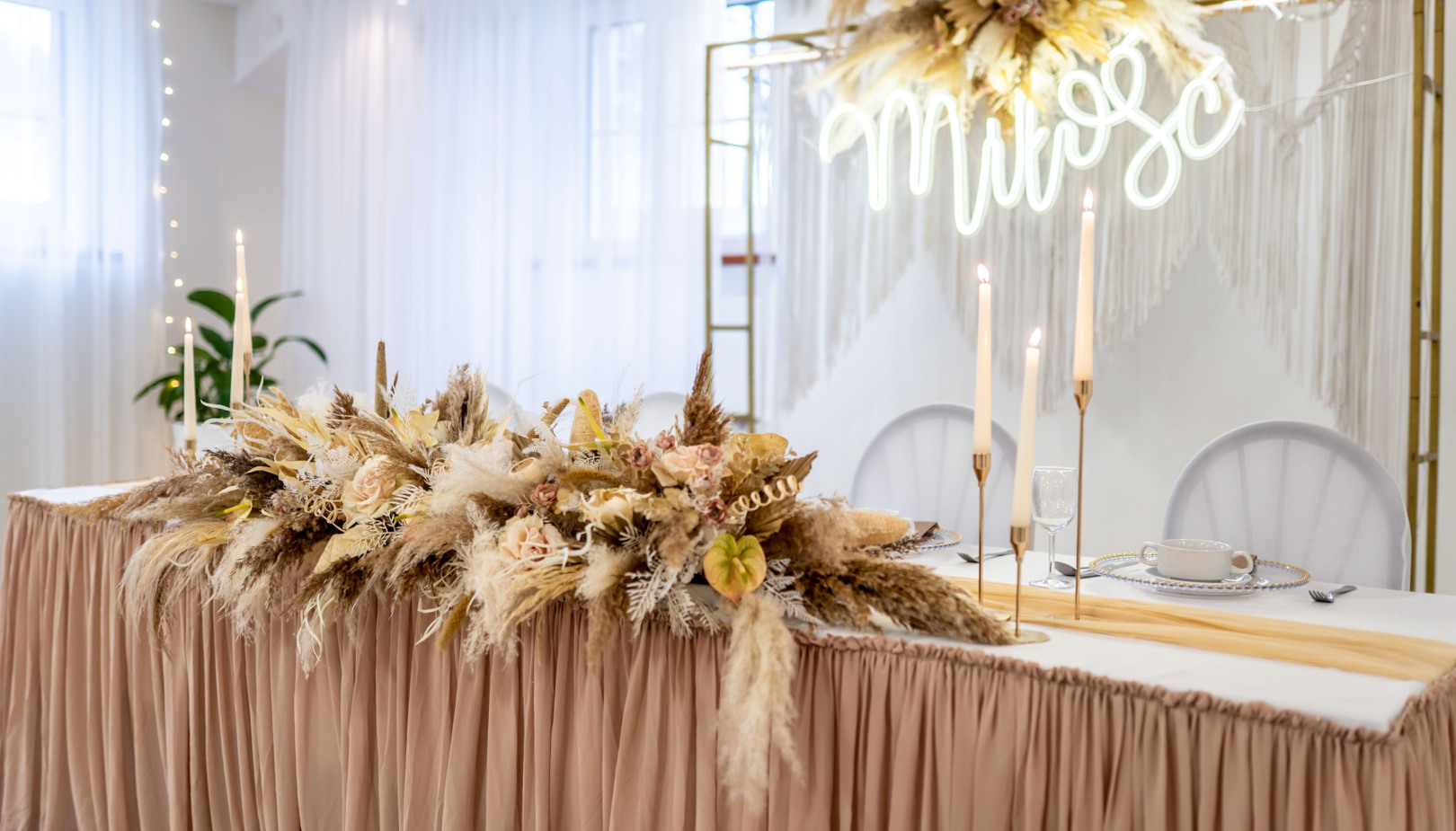 Wedding head table with blush-beige draped skirt, pampas grass centerpiece, dried florals, tall candles, place settings and neon 'miłość'