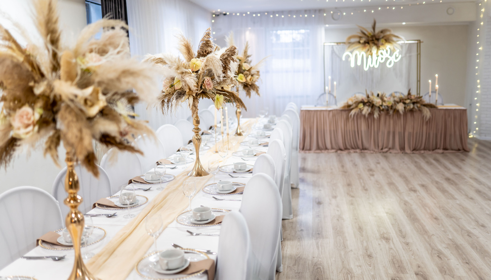 Elegant wedding banquet: long white-covered tables, gold pampas-grass centerpieces, warm fairy lights, decorated head table.