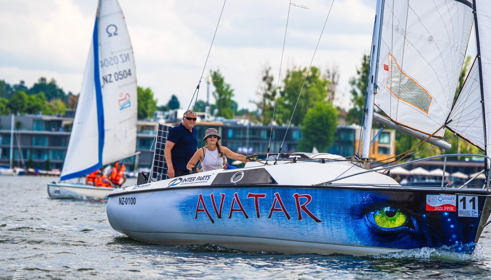 Para żegluje jachtem „Avatar” z niebieską grafiką oka obok hoteli nadbrzeżnych — relaksujący rejs po marinie.