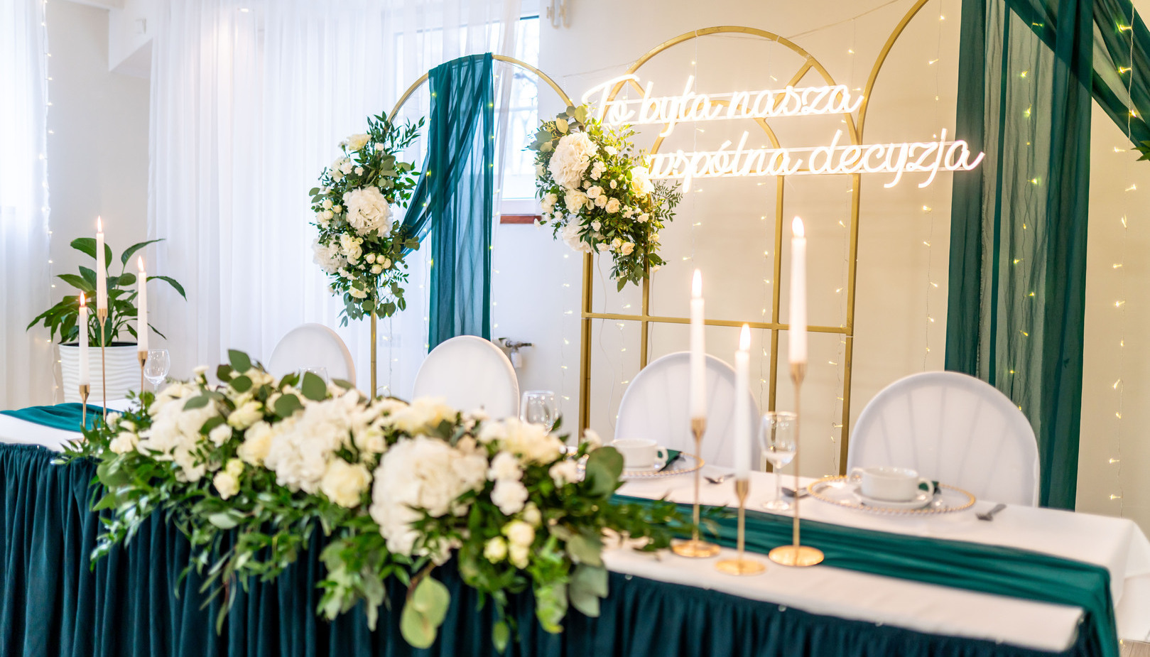 Elegant wedding head table with deep emerald drapery, white floral garland, gold arches, glowing neon sign and tall candle centerpieces.