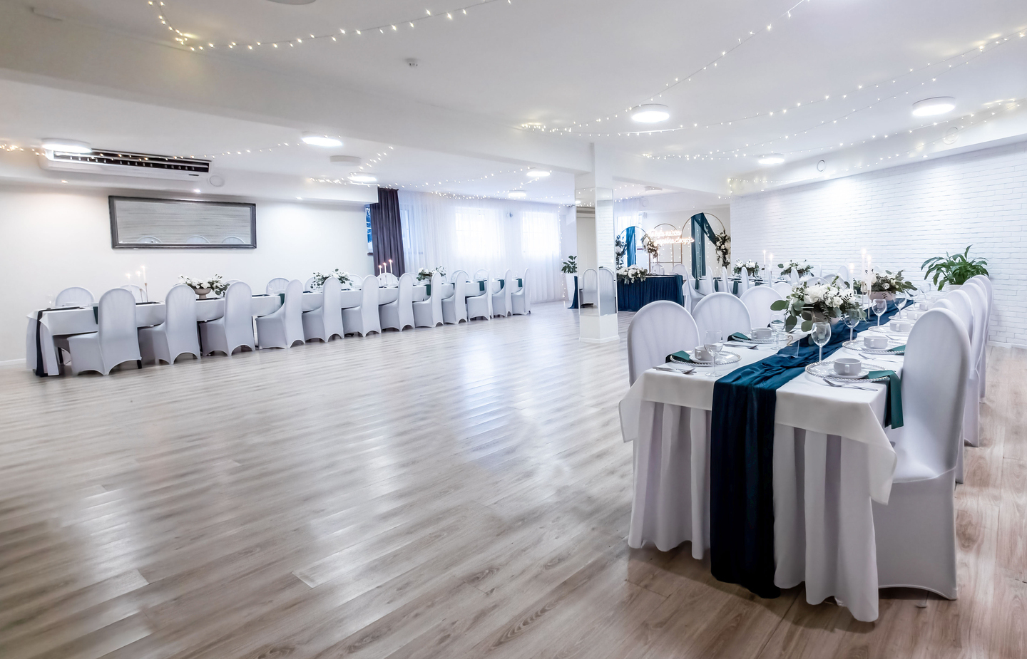 Bright modern banquet hall set for a wedding with long white-draped tables, teal runners, floral centerpieces, chair covers and fairy lights