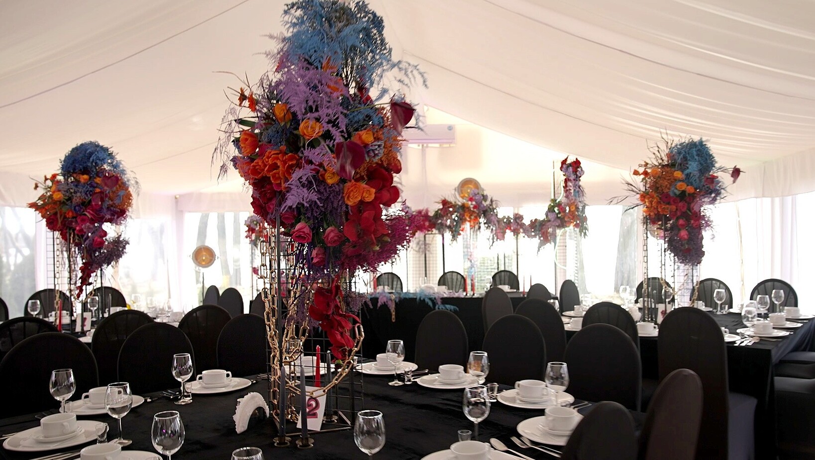 Festzelt mit schwarzen Tischdecken, gedeckten Tischen und hohen, bunten Blumenarrangements für einen Empfang