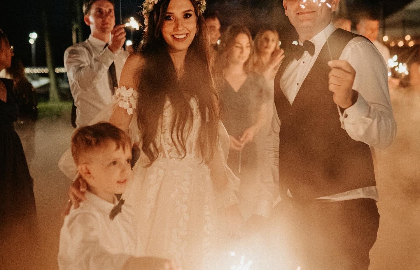 Panna młoda w wianku i koronkowej sukni i pan młody w smokingu uśmiechają się na wieczornym plenerowym weselu, goście i dziecko trzymają zimne ognie.