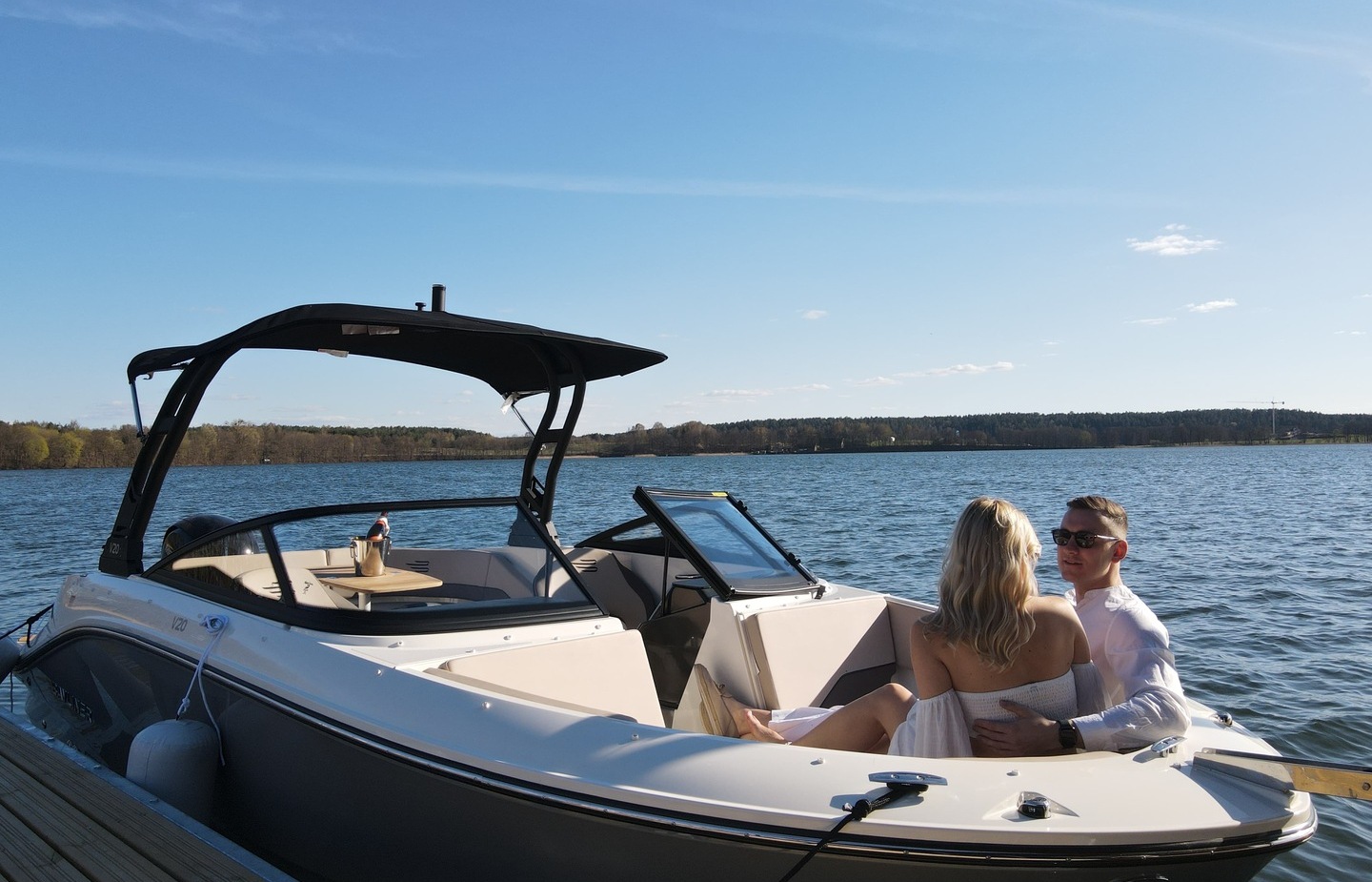 Paar entspannt auf modernem Motorboot am Steg, überdachte Sitze, Champagner-Eimer, ruhiger See mit Baumreihe