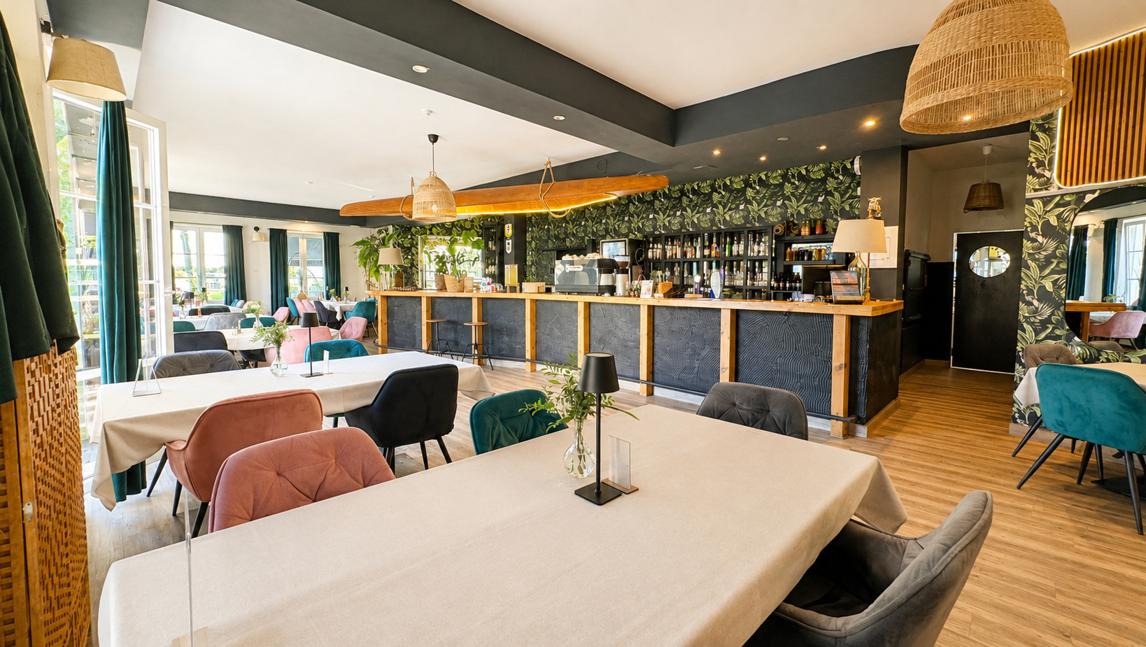 Jasna restauracja hotelowa z drewnianym barem, tropikalną tapetą, rattanowymi lampami, aksamitnymi różowymi i turkusowymi fotelami.