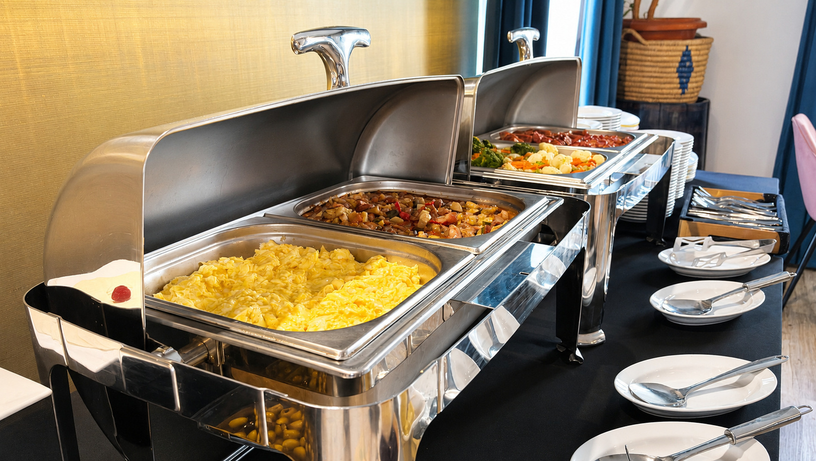 Frühstücksbuffet mit Edelstahl-Warmhaltern: Rührei, gemischtes Gemüse und Eintöpfe; gestapelte Teller und Servierlöffel.