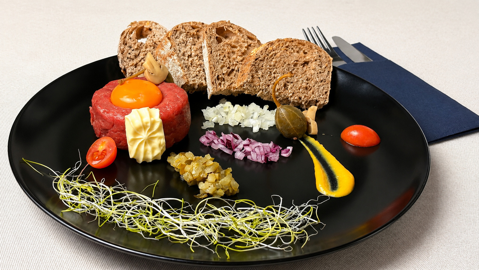 Gourmet beef tartare topped with raw egg yolk, butter, diced onions, pickles and caperberry, sprouts and multigrain bread on a black plate.