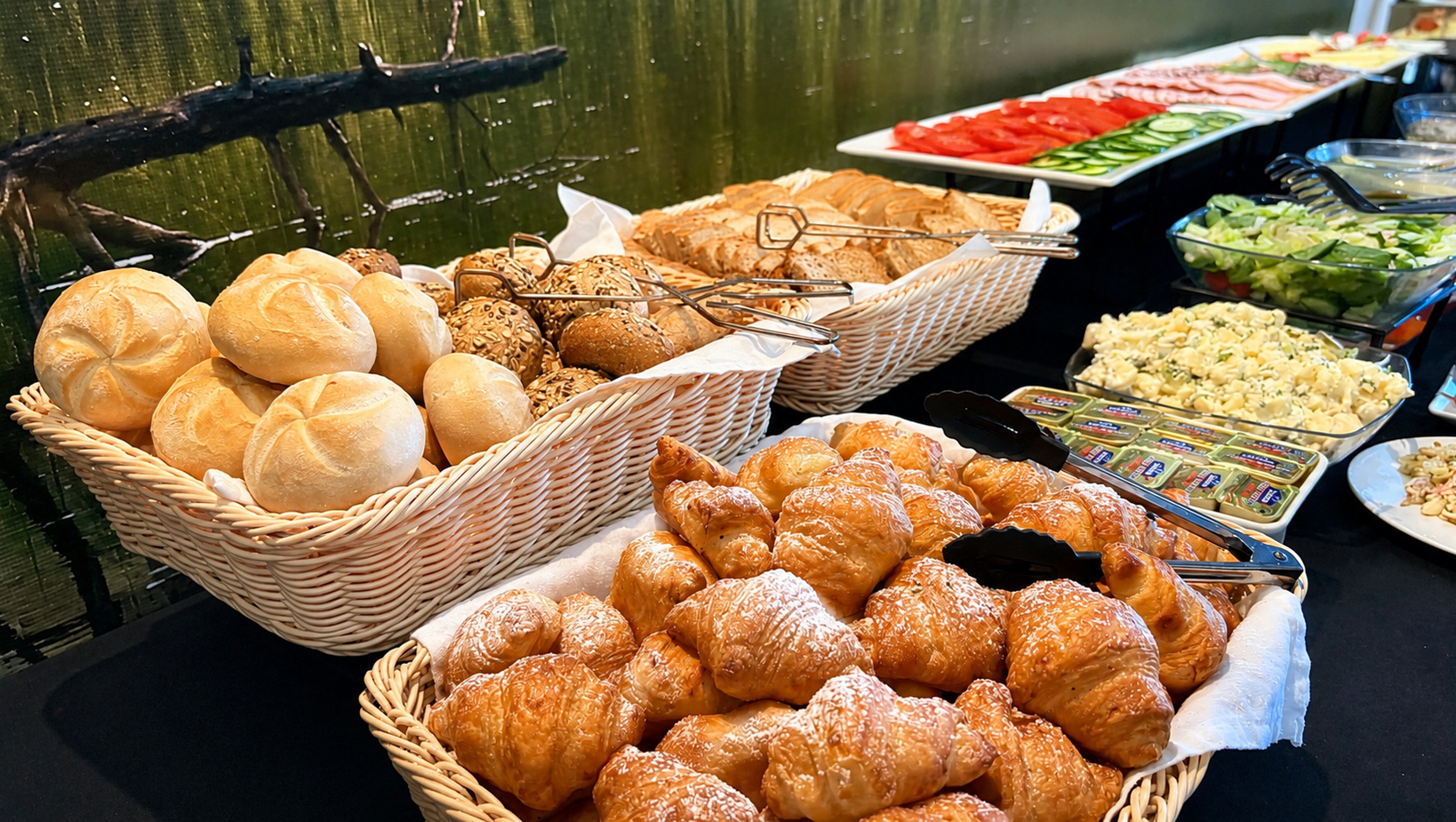 Frühstücksbuffet: Körbe mit frischen Brötchen, Körnerrollen und Puder-Croissants; dazu Salate, Tomaten- und Gurkenscheiben und Aufstriche.