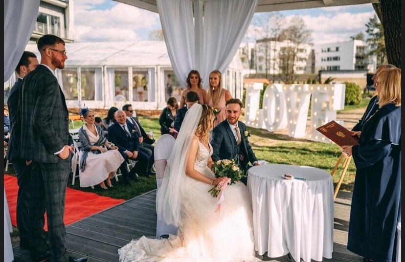 Plenerowy ślub pod białą altaną na terenie hotelu, panna młoda w sukni, pan młody przy stole do podpisów, siedzący goście i świetlny napis 'LOVE'.