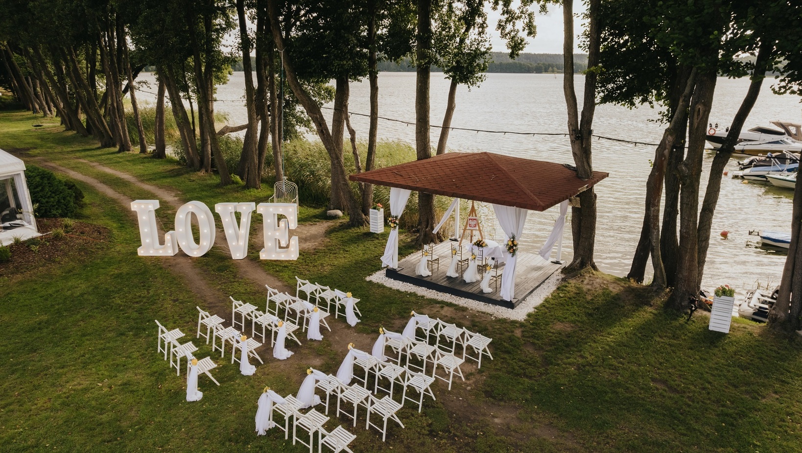 Plenerowa ceremonia nad jeziorem: altana z białymi zasłonami i kwiatami, rzędami krzeseł i świecącymi literami LOVE.