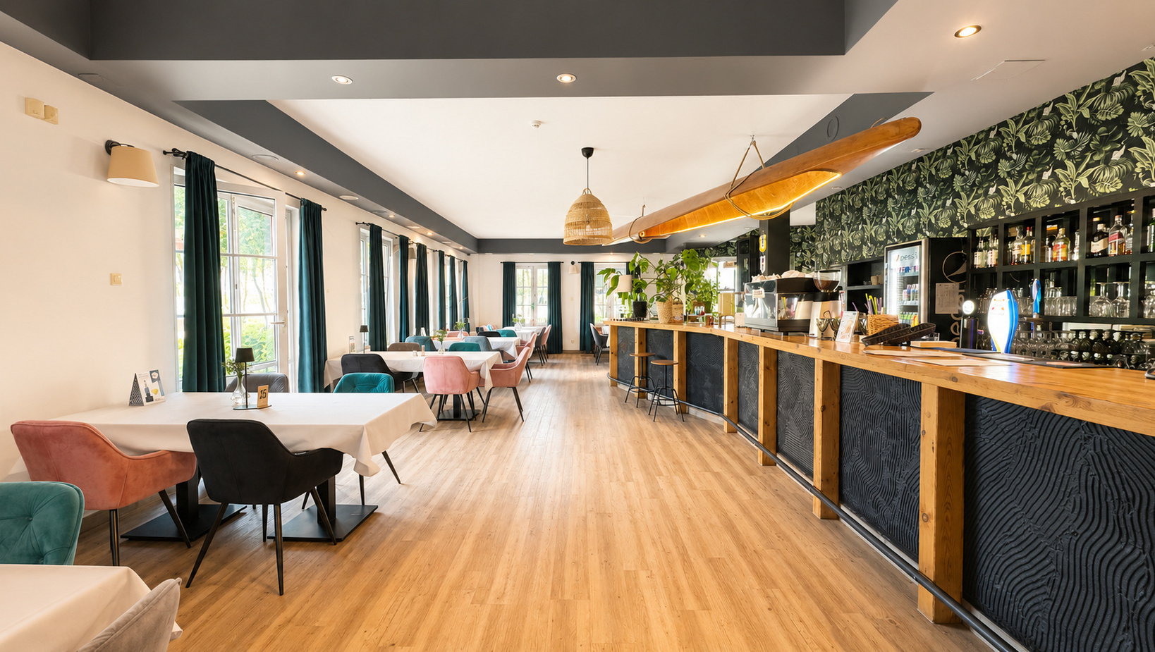 Helles Hotelrestaurant mit langer Holzbar, tropischer Tapete, türkisen und rosafarbenen Stühlen, großen Fenstern und Pflanzen.