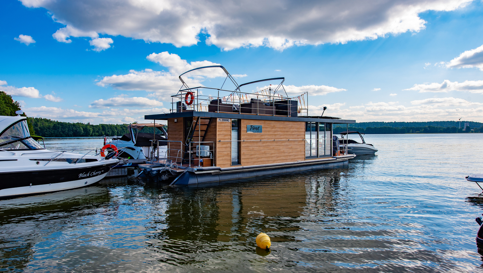 Holzhausboot mit Dachterrasse und Loungemöbeln, Glastür und Rettungsring, neben Yachten an einem ruhigen See vertäut.