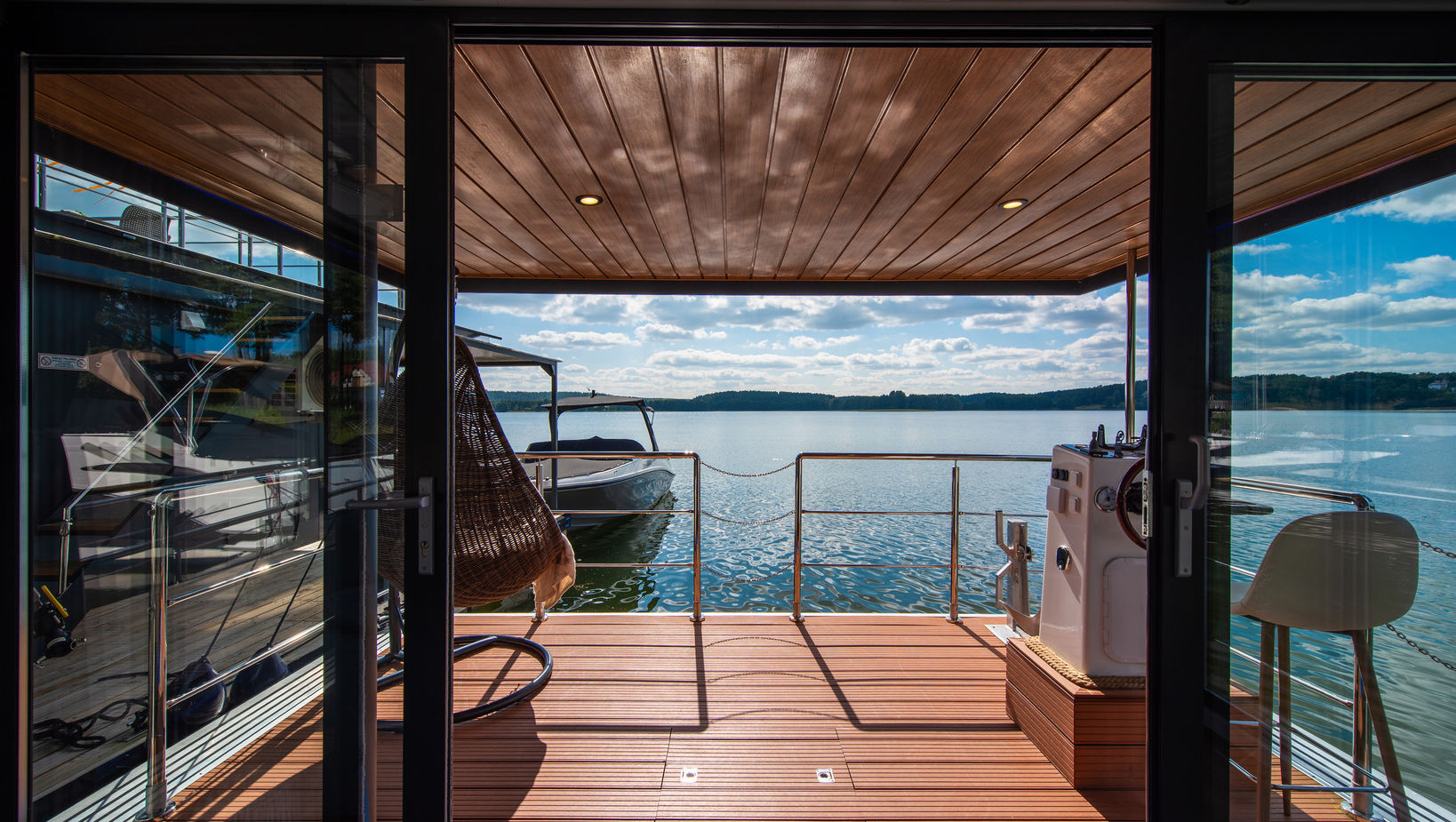 Überdachte Holzterrasse mit Blick auf ruhigen See, angelegtem Boot, hängendem Korbsessel und Metallgeländer.