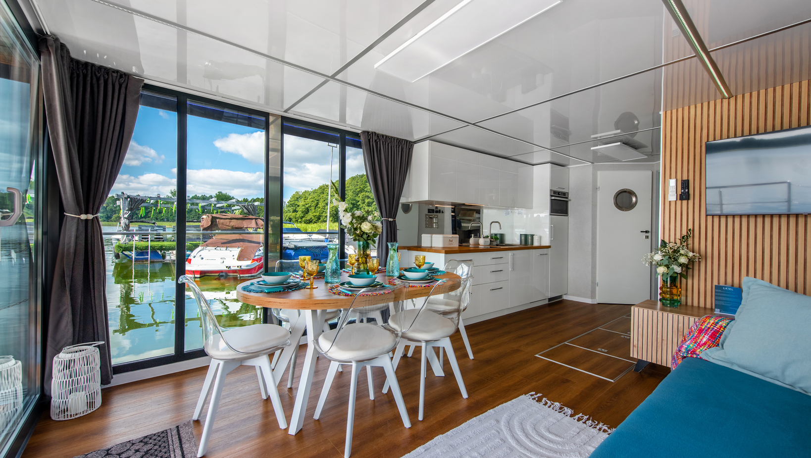 Waterfront suite with floor-to-ceiling glass, marina view, set dining table, compact white kitchen, wooden accents and cozy lounge.