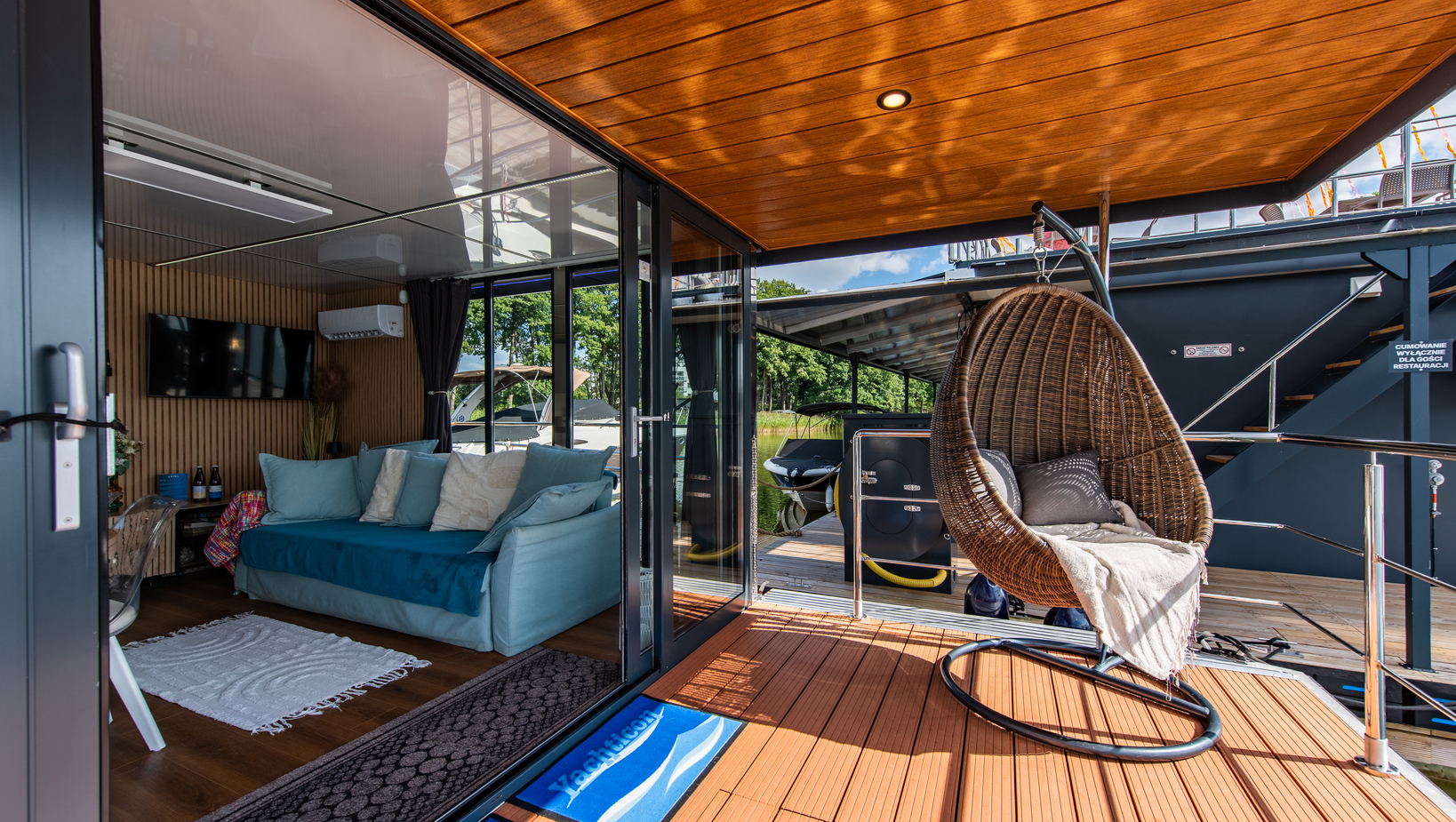 Private waterfront deck with wooden ceiling, hanging rattan chair and glass doors to a cozy lounge with sofa and TV.