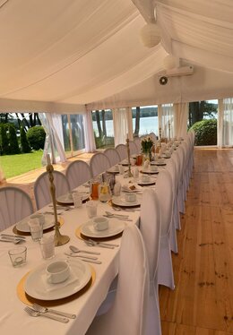 Lakeside white marquee set for a banquet: long table with white linens, gold chargers, candles, pitchers, wooden floor and lake view.