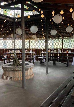 Covered wooden pavilion set for a reception with paper lanterns and string lights, central brick firepit, long benches and buffet tables.