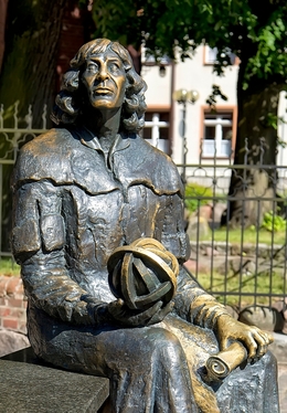 Bronzefigur sitzend, hält eine Armillarsphäre und eine Schriftrolle, vor einem schmiedeeisernen Zaun und historischem Gebäude