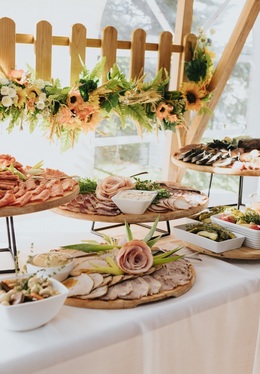 Elegantes Buffet mit Aufschnitt, geräuchertem Fisch und Salaten auf Holzplatten, geschmückt mit Sonnenblumen-Girlande in hellem Zelt