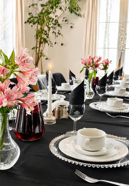 Elegant dining table with black linens, beaded chargers, white china, black napkins in glasses, pink flowers and lit candle.