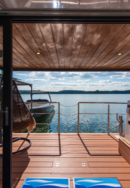 Covered wooden terrace overlooking a calm lake with a moored boat, wicker hanging chair and metal railing.