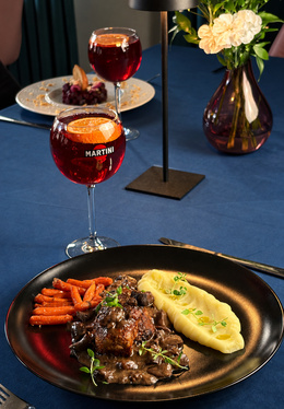 Gebratenes Fleisch mit Pilzsauce, cremiges Kartoffelpüree und glasierte Möhren auf schwarzem Teller; Martini-Aperitif im Stielglas, Lampe und Vase