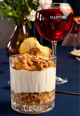 Schichtdessert aus Joghurt mit Granola und getrockneter Apfelscheibe, daneben ein großes rotes Martini-Glas und weiße Blumen auf dunkelblauem Tisch.