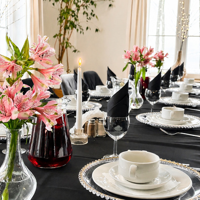 Eleganter Tisch mit schwarzem Tuch, Perlendekor-Tellern, weißem Geschirr, schwarzen Servietten, Blumen und brennender Kerze.
