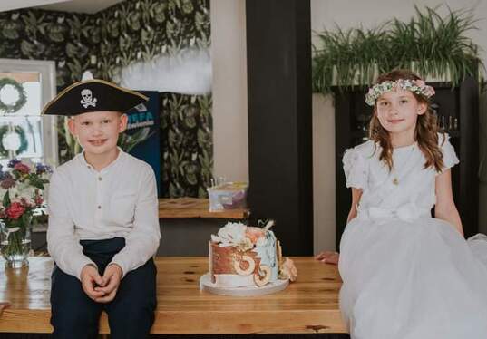 Two children in a party room — boy in a pirate hat and girl in a white dress with flower crown seated by a decorated cake on a wooden table