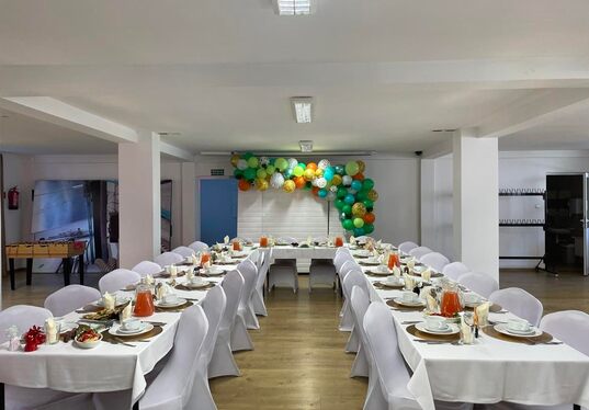 Elegant private event room with U-shaped tables, white chair covers, place settings and pitchers of orange drink, green-yellow balloon arch at back